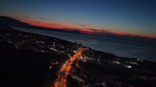 Aerial drone view of a lit coastal road winding through a seaside town under a fading red sunset sky, Rethymno, Greece