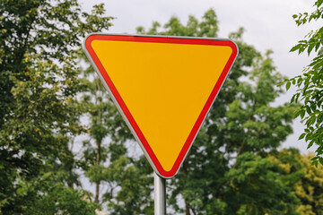 Obraz premium Blank yield sign stands outdoors. Yellow triangle has red border. Green trees create natural background. Empty road sign allows customization. Traffic concept supports safety designs.