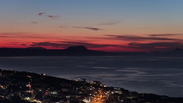Aerial drone view of a coastal town glowing at night with a lit harbor beside the dark sea, Rethymno, Greece