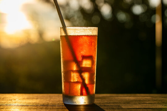Refreshing iced tea cocktail in glass at golden hour, closeup. Cold iced tea or cocktail with ice cubes and a straw in misty glass on wooden table outdoors during sunset golden hour at summer