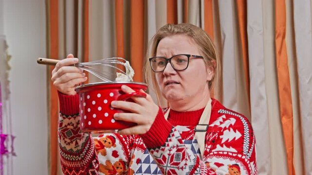 A woman in a christmas sweater and apron holds a red polka dot pot at home, smiling at first then frowning as she whisks a failed dessert with a wire whisk.