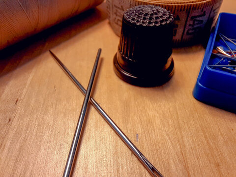 Both crossed metal sewing needles rest on a wooden table next to a black thimble. A measuring tape, thread, and blue box of pins blur in the warm background of this close-up tailoring crafts workshop.