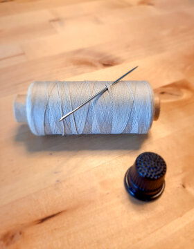 A spool of gray sewing thread with a metal needle tucked in sits on a wooden table next to a dark thimble. A close-up view of tailoring and handicraft equipment for mending, repair, and diy crafts.