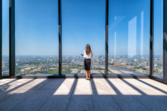Rear view of a business woman looking out of her large office windows high above the urban skyline of the City of London, England