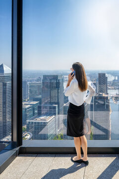 A business woman talking on the phone looks at the skyline of the financial district Canary Wharf in London from her office window