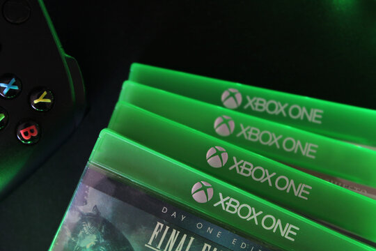 Xbox One game cases stacked with controller background. Editorial macro shot of green video game boxes spine and gamepad buttons with atmospheric lighting. Close-up view.