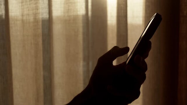 Excessive phone use silhouette: backlit hand holding smartphone and scrolling near window curtains