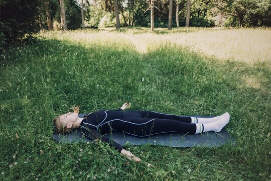 Woman lies on exercise mat in grass with arms spread wide during outdoor practice. Somatic yoga practice, nervous system regulation, trauma informed yoga.