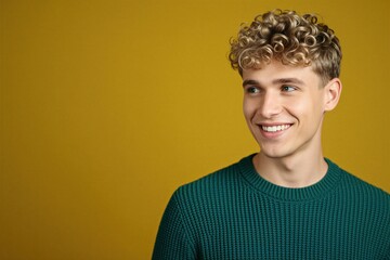 Fototapeta premium Young blond man with curly hair wears green sweater. He smiles looking to the side against a yellow background. Natural expression, confident and happy.