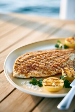 Grilled fish fillet with charred lemon and fresh herbs served on a wooden table by the sea at golden hour