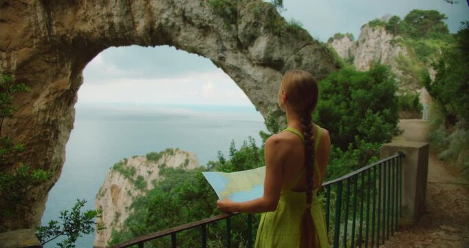 A woman studies a map while admiring Capri Natural Arch overlooking the calm sea in overcast summer day