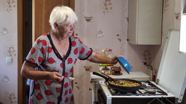 A gray-haired old woman cooks vegetable stew in a frying pan on a gas stove. Poverty, destitution. Social inequality. A small pension.