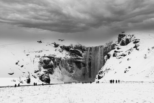 Tourists at Skogafoss waterfall, snowy and icy rock face, dark clouds above, black and white photo, Sudurland, Iceland