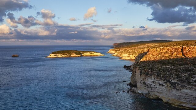 Wide angle, morning light, sunrise, cloudy blue sky, dark sea, cliff illuminated by morning light, Isola dei Conigli, Spiaggia dei Conigli, Riserva Naturale Orientata Isola di Lampedusa, Lampedusa Island, Agrigento Province, Pelagic Islands, Sicily, Italy