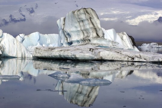 Iceberg with black stripes reflected in the water, mountains rising out of the mist, J&ouml;kulsarlon, glacier lagoon, Scandinavia, Iceland
