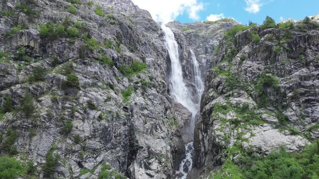 Aerial drone rises from a rocky gorge with Shdugra waterfall, opening a panoramic view of snow-capped Mount Ushba, the river below, and a blue sky with scattered clouds. Original 4K footage.