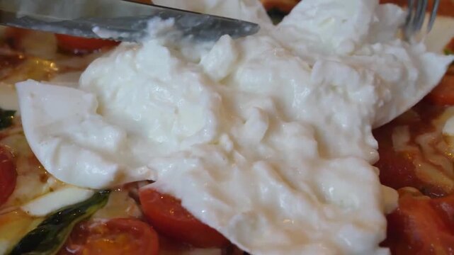 close-up shot of a knife and fork slicing into a large ball of creamy burrata cheese at the center of a fresh Italian pizza. The soft, rich interior spills over cherry tomatoes and basil, showcasing a