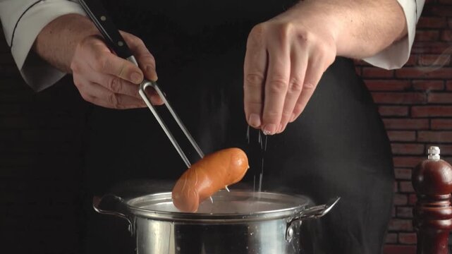 Chef in black apron lifts sausage from pot of boiling water with fork and salt, steam rising around it, standing near rustic brick wall in kitchen