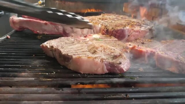 High-quality footage of premium beef cuts, including Tomahawk and T-bone steaks, sizzling on a barbecue grill. The scene captures detailed grill marks, rising smoke, and dancing flames over hot coals,