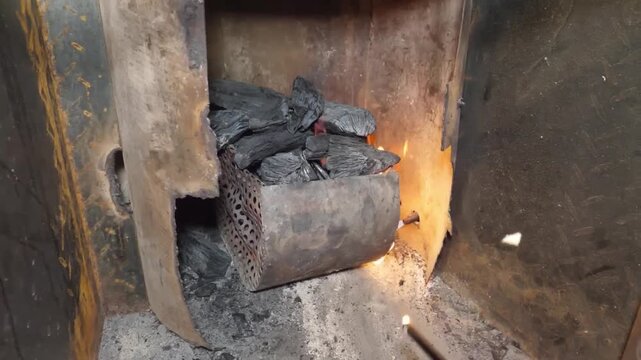 A detailed shot showing the initial process of lighting charcoal in a metal grill. Vibrant orange flames flicker around dark, textured charcoal pieces, creating a dramatic atmosphere for outdoor cooki