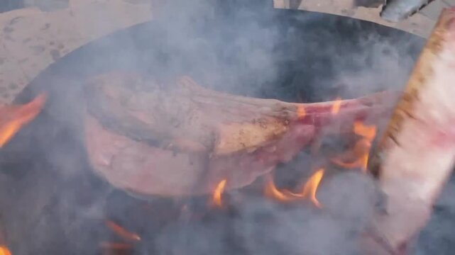 Two premium beef tomahawk ribeye steaks being grilled over an open fire on a flat barbecue surface. The footage captures vibrant orange flames, rising smoke, and the searing process of high-quality ma