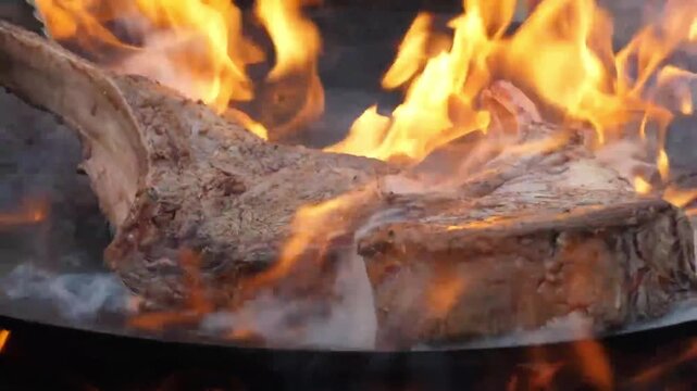High-angle shot of two large, bone-in tomahawk beef steaks grilling over an open fire. The footage captures vibrant orange flames and thick white smoke swirling around the meat as it develops a charre