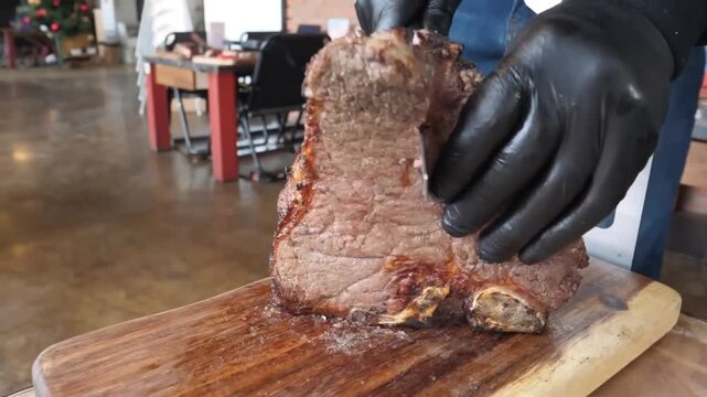 A professional chef wearing black gloves carefully carves a large, juicy, perfectly grilled medium-rare beef steak on a rustic wooden cutting board. The close-up shot highlights the rich texture, pink