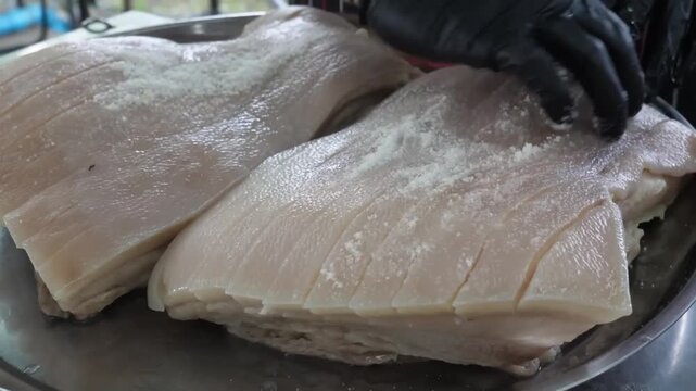 Close-up footage of a hand in a black glove sprinkling salt over two large, scored slabs of raw pork belly on a metal tray, preparing for a crispy barbecue or oven-roasted crackling finish.