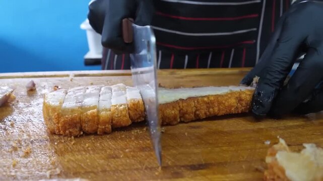 Close-up footage of a person slicing a large piece of freshly roasted pork belly with a sharp knife on a wooden board. The scene highlights the crunchy, golden-brown crackling skin and the succulent, 