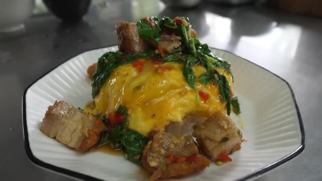 A vibrant close-up shot of authentic Thai Pad Kra Pao Moo Krob, featuring crispy pork belly stir-fried with chili, garlic, and fragrant holy basil, topped onto a soft, creamy Thai-style omelet and ste