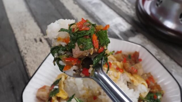 A close-up shot of a spoon lifting a portion of authentic Thai Pad Kra Pao, featuring savory stir-fried meat with fresh holy basil leaves and vibrant red chilies, served over a bed of steamed jasmine 
