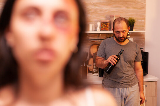 Abusive husband drinks alcohol while looking toward his traumatized wife, connecting domestic violence, injury and fear in a kitchen scene.