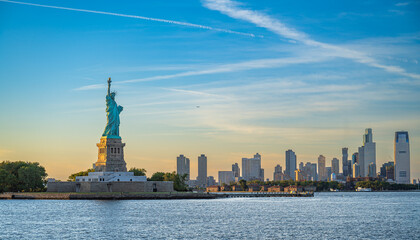 Statue of Liberty beside Jersey City skyline, New York. Copper monument and waterfront towers stand across Upper New York Bay under blue evening sky. © Vadim