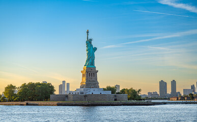 Statue of Liberty beside Jersey City skyline, New York. Copper monument and waterfront towers stand across Upper New York Bay under blue evening sky. © Vadim