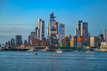 Fototapeta na wymiar Hudson Yards skyline across Hudson River at dusk. Glass skyscrapers rise above waterfront piers as boats cross the river under a blue evening sky.