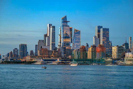 Hudson Yards skyline across Hudson River at dusk. Glass skyscrapers rise above waterfront piers as boats cross the river under a blue evening sky.