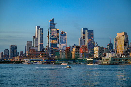 Hudson Yards skyline and motorboat across Hudson River. Illuminated glass towers rise behind waterfront piers as a small vessel crosses blue hour water.