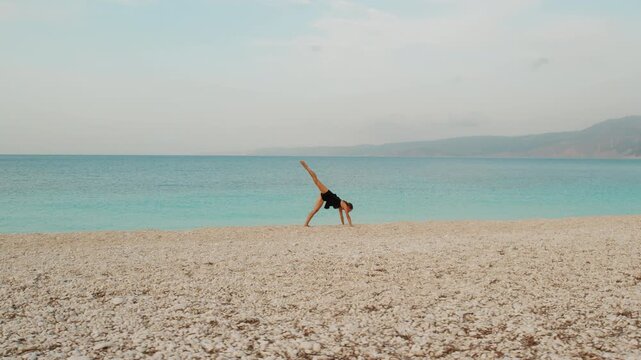 Woman practices yoga pose on wide pebble beach beside calm turquoise summer sea