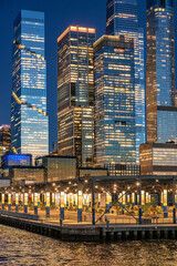 Fototapeta premium Pier 76 beneath illuminated Hudson Yards office towers. Covered waterfront structure with lit columns borders the river below dense glass skyscrapers at night.
