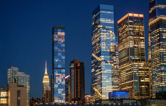 Empire State Building framed by Hudson Yards towers at night. Wide Manhattan view combines historic landmark, modern glass high rises, and The Shed near the base.