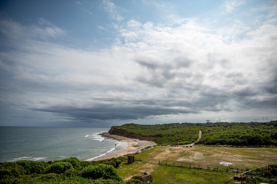 Montauk coastal landscape, ocean cliffs and cloudy sky. Atlantic shoreline curves around an eroded green bluff beside an open field under mixed clouds.