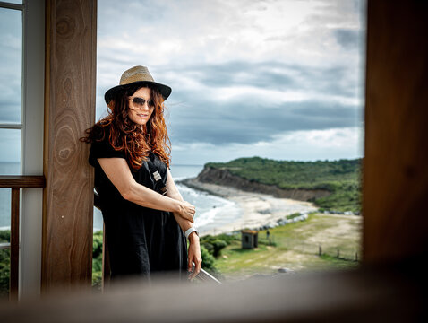 Female tourist poses near ocean viewpoint on Long Island. Medium portrait shows a woman in hat and sunglasses beside timber framing with a blurred shoreline behind.
