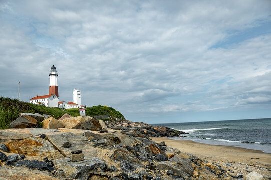 Montauk Point Lighthouse above rocky Atlantic shoreline. Low coastal view includes the tower, white buildings, boulders, sandy beach, waves, and cloud cover.