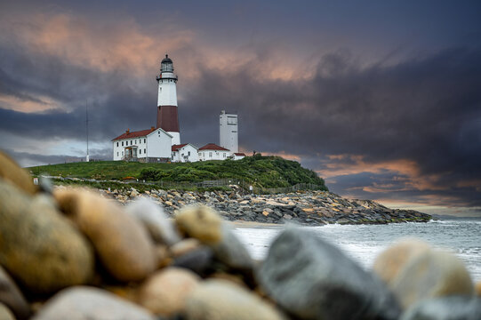 Montauk Point Lighthouse beneath dark evening storm clouds. Historic tower and white buildings stand above a rocky shoreline with blurred stones and breaking waves.