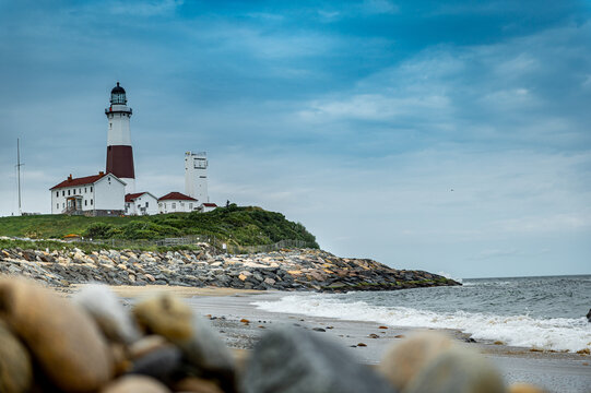 Montauk Point Lighthouse overlooking Atlantic beach in New York. Coastal complex rises above a rocky headland with surf, wet sand, foreground boulders, and blue cloud cover.