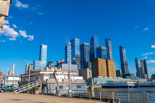 Passenger vessels dock below Hudson Yards skyline, Manhattan. Waterfront view includes glass towers, pier structures, railings, blue sky, and the Empire State Building.