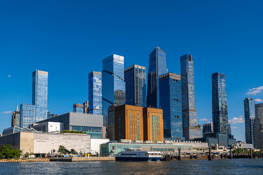 Hudson Yards skyline rises above Manhattan waterfront. Glass towers, low pier buildings, a moored ferry, and the daytime moon appear beneath clear blue sky.