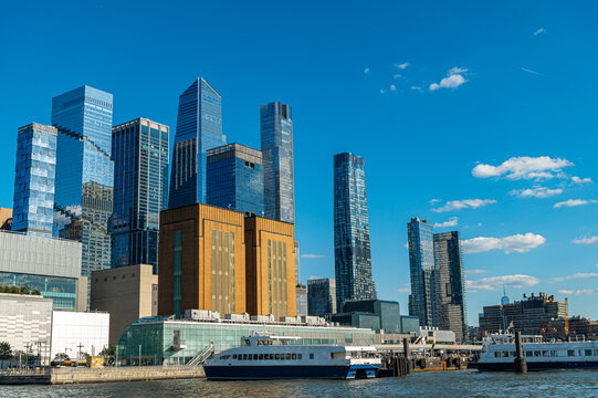 Ferries dock beneath Hudson Yards skyscrapers in Manhattan. Passenger vessels line the waterfront below glass towers and scattered clouds in daylight.