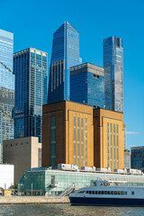 Fototapeta premium Hudson Yards glass towers behind Manhattan waterfront building. Vertical composition shows layered skyscrapers, brick architecture, a ferry, and clear blue sky.