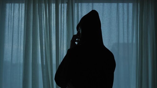 Silhouette of a mysterious hooded man making a phone call at night in dark room with curtains. Shady business deal by hacker or spy. Secured communication by phone call